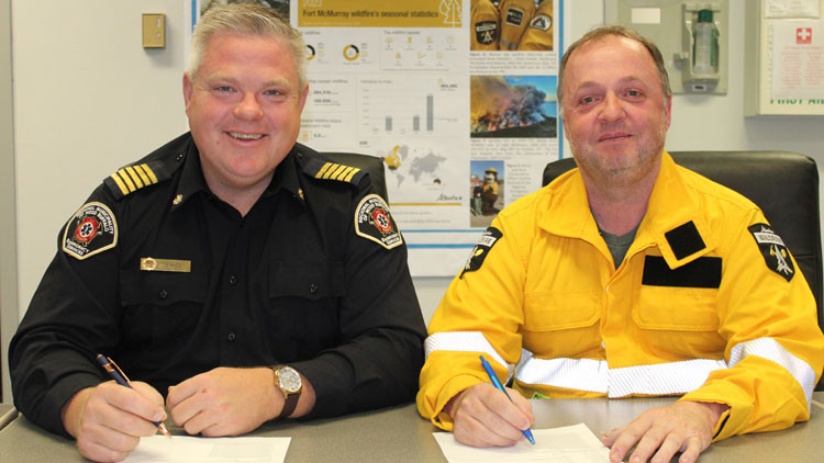 Jody Butz Wendell Pozniak - a man in a black uniform smiling at the camera with a pen in his hand signing a document, sitting next to a man in a yellow uniform with a pen in his hand signing a document