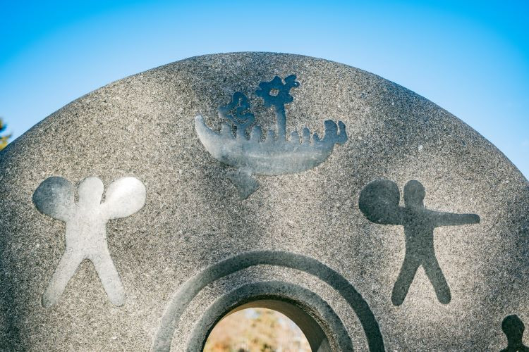Photo of Petroglyphs on the residential school monument, first depicting a person holding two bundles, then a ship, then a person holding one bundle.