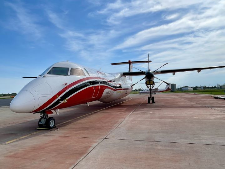 A red, white, and black propeller airplane is parked on a concrete runway under a partly cloudy sky. The runway extends into the distance with green grass visible on the right side.