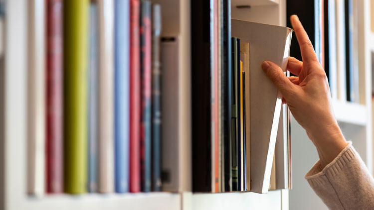 A person's hand taking a book of a shelf with multiple books
