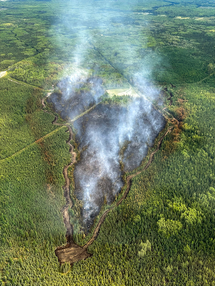 Aerial view of a contained wildfire in a dense forest. Light smoke rises from a blackened burn area surrounded by green trees, with a dozer guard encircling the fire and a cleared access point visible at the bottom of the image.
