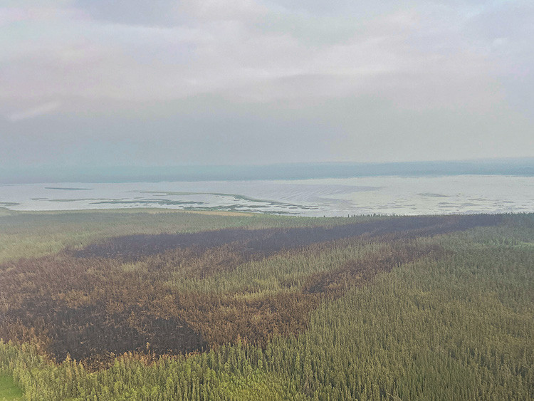 Aerial view of a forested landscape showing a large burned area stretching toward wetlands and open water. Hazy skies and smoke reduce visibility, highlighting the scale of recent wildfire activity across the region.