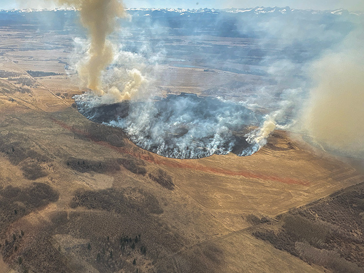 Aerial view of a large wildfire burning across open grassland. A wide, dark burn area is ringed by active flames, with dense white and tan smoke rising in several plumes toward distant mountains under a clear sky.