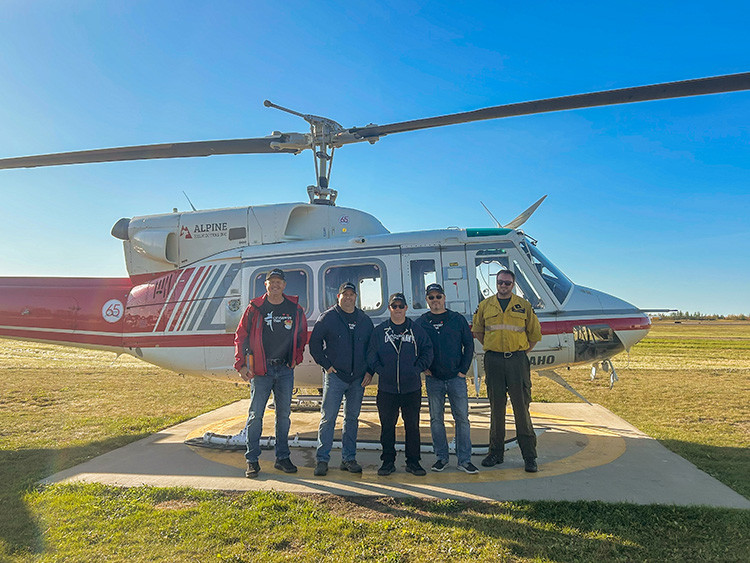 The winners from the 'Take the Pledge Campaign' pose for a photo with a pilot and Alberta Wildfire personnel.  They stand in front of  a parked red and white helicopter with its rotor blades visible overhead. 