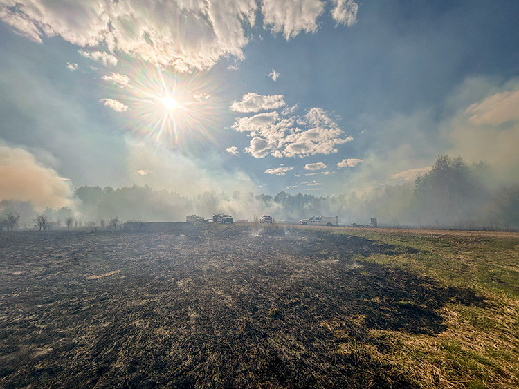 Charred field in foreground under bright sun and scattered clouds, with light smoke draping a tree line. Several emergency vehicles with lights on are positioned along the edge of the burned area during wildfire response.