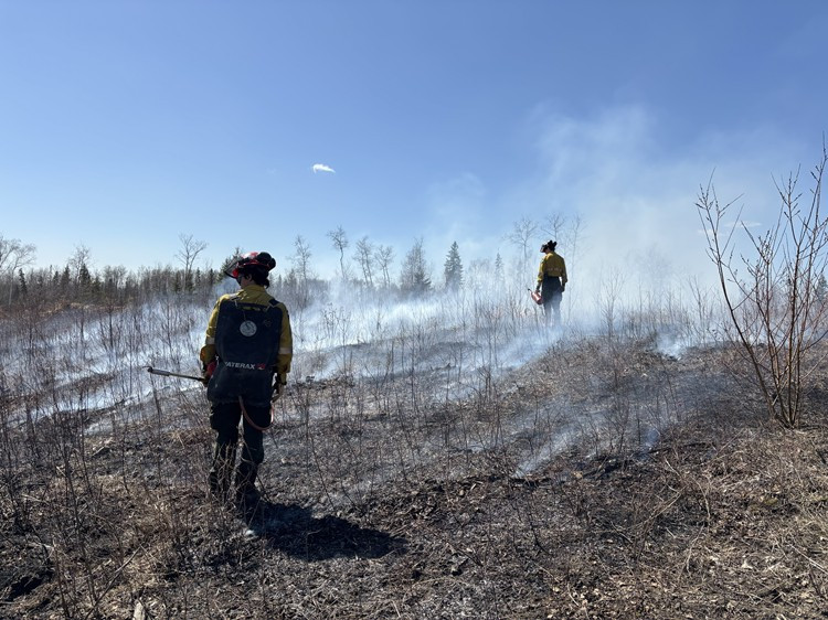 Firefighters in yellow protective gear use drip torches and portable fire pumps to manage a controlled burn in a hilly area surrounded by thick, fog-like smoke under a clear blue sky.