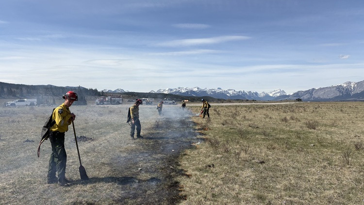 Firefighters in yellow protective gear use hand tools and drip torches to manage a controlled burn across a dry grass field; smoke drifts along a line of small flames, with fire trucks and snow-capped mountains visible in the background.