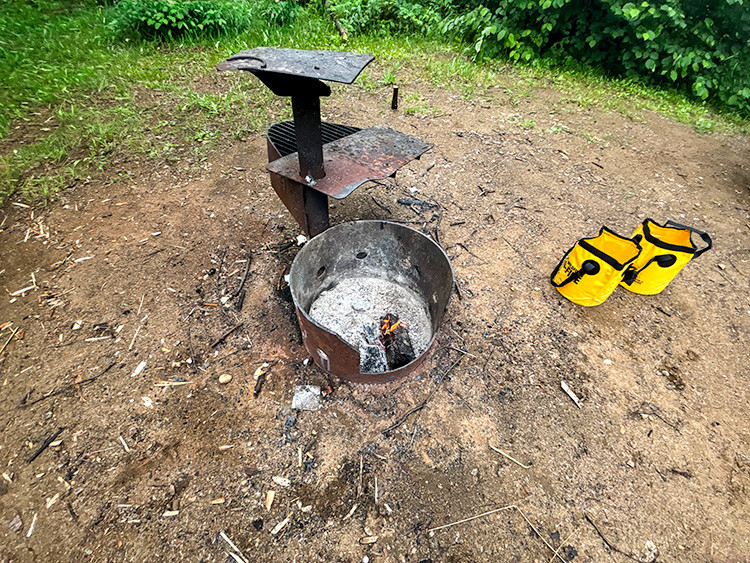 Metal fire ring and grill at a campsite with a small smouldering ember in the ash. Two yellow collapsible water buckets sit nearby on bare ground, surrounded by green vegetation.