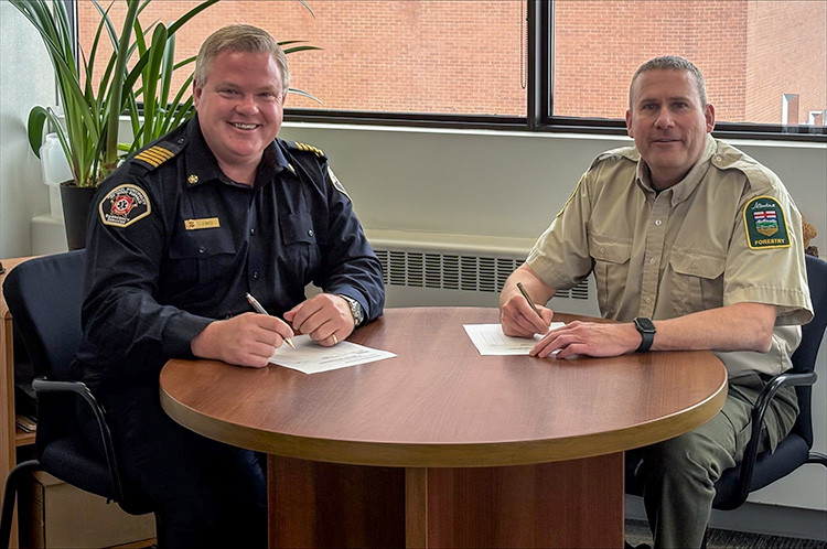 Two uniformed officials sit at a round table indoors, each signing documents. One wears a dark fire service uniform, the other a tan forestry uniform. A large window and potted plant are visible behind them.