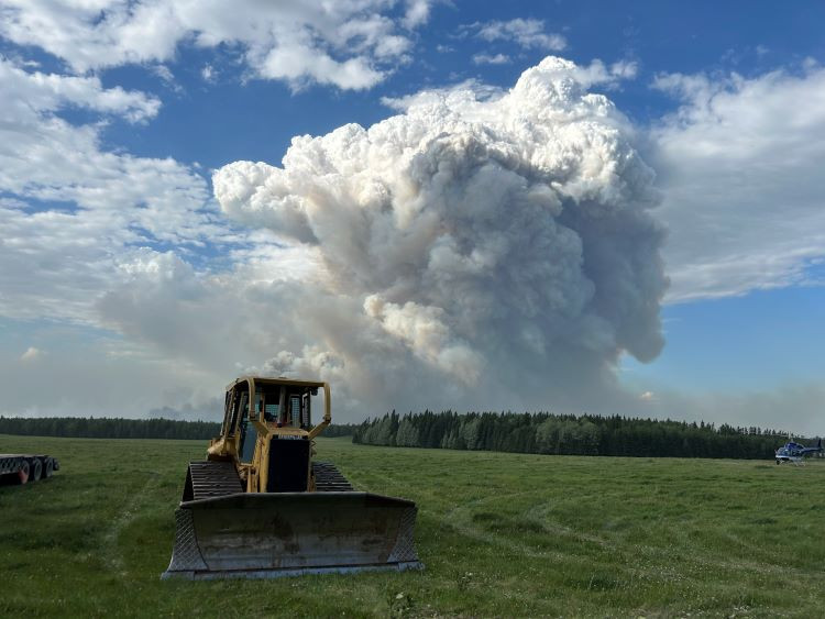 Dozer and a helicopter is in front of a large column of smoke.