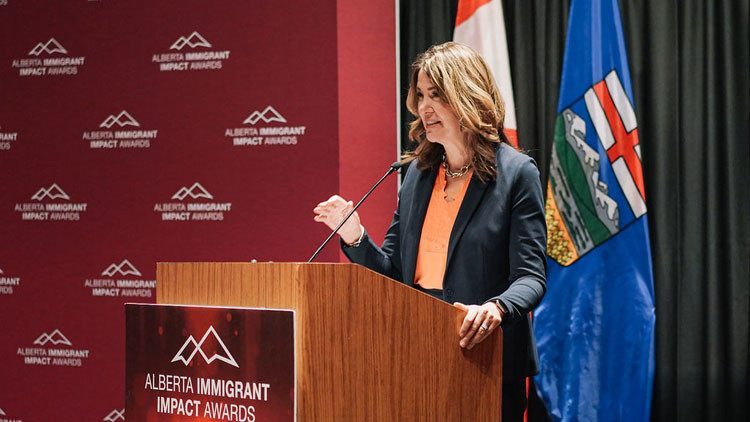 Premier Smith speaking at the podium on stage at the 2025 Alberta Immigrant Impact Awards ceremony