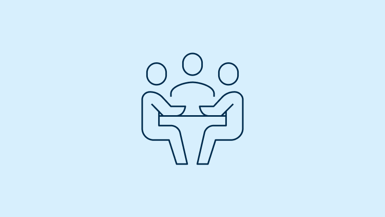 Icon of 3 people seated at a desk having a meeting.