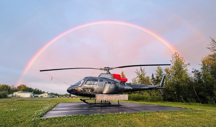 A black helicopter sits on a grassy helipad under a vivid rainbow arching across a pastel evening sky. Trees and small buildings are visible in the background.
