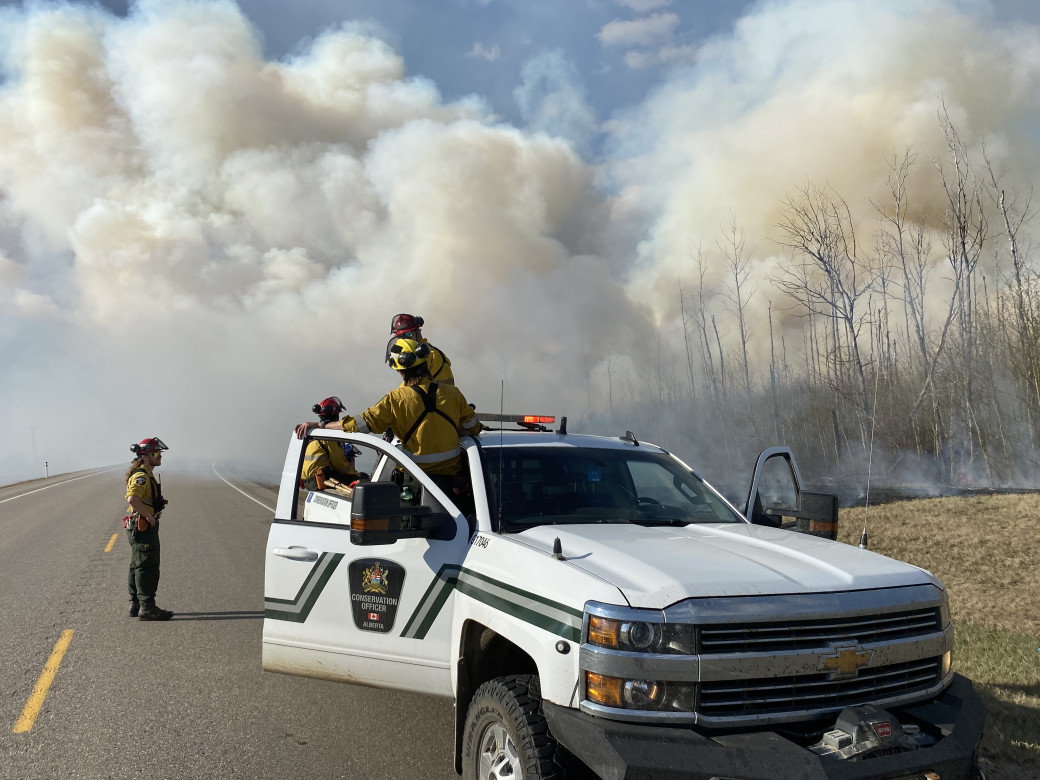 A crew of wildland firefighters exit a conservation officer's patrol truck. A plume of smoke rises from the forest in the distance