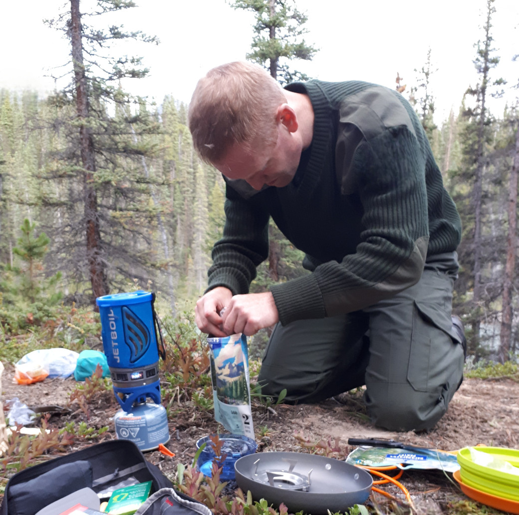 A young male camper uses his propane stove to boil water for his dehydrated meal. He is in the trees and it's an overcast day. 