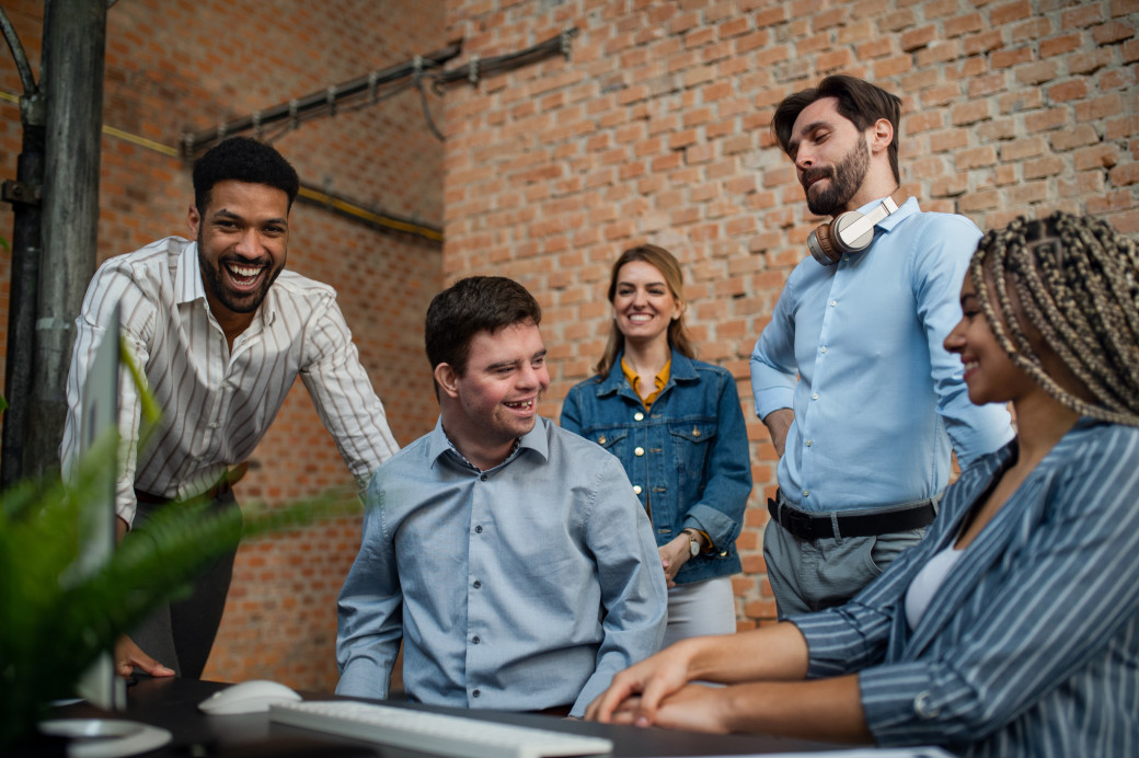 Group of people happily and assist a person with developmental issues with work on a computer.