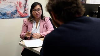 Woman sitting at desk speaking to a man. Woman has a pen in her hand with papers on the desk