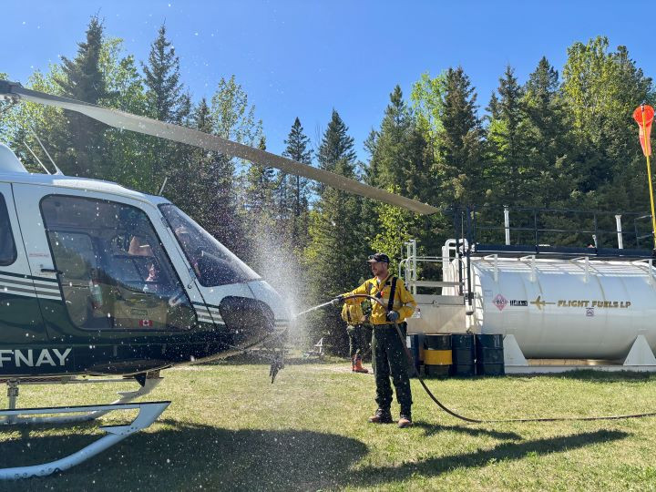 With clear clue skies and forest in the background, a wildland firefighter sprays water from a fire hose on to the front of a helicopter.