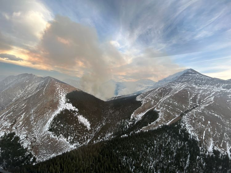 A mountain valley, with patches of snow on the slopes and dense evergreen forest in the lower areas. A column of smoke rises from the centre of the valley, spreading into the sky, indicating an active wildfire. The sky is partly cloudy with soft sunset tones.