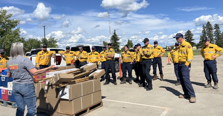 A group of firefighters wearing yellow wildfire uniforms stand together in a parking lot, listening to a woman in a grey shirt who is speaking to them. Several cardboard boxes and supplies are stacked on a wooden pallet in front of her. The sky is blue with scattered clouds.