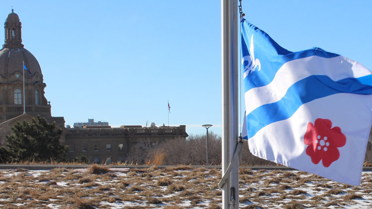 Alberta legislature building with Franco-Albertan flag on a pole in the foreground: white flag with a white fleur-de-lys on the top left blue corner, diagonal blue band across the centre, and red wild rose on the bottom right white corner