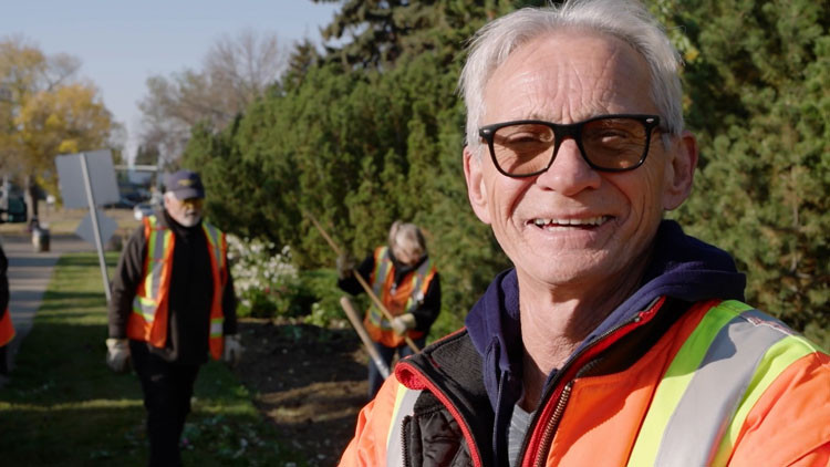 Doug stands in the foreground, wearing an orange and yellow reflective work jacket and smiles at the camera. Two workers are behind him working with some trees.