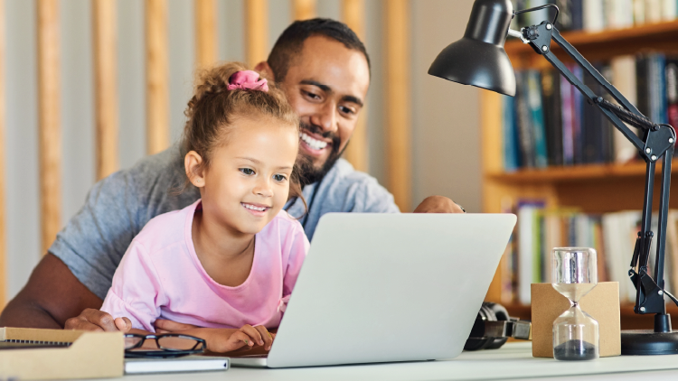 A father sitting at a computer with his young daughter, both looking at the screen and smiling.