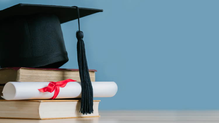 Black graduation cap sitting on two books with a diploma rolled up tied in a red ribbon