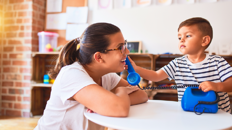 A boy toddler at a small round table holding a toy phone receiver to a young woman's ear
