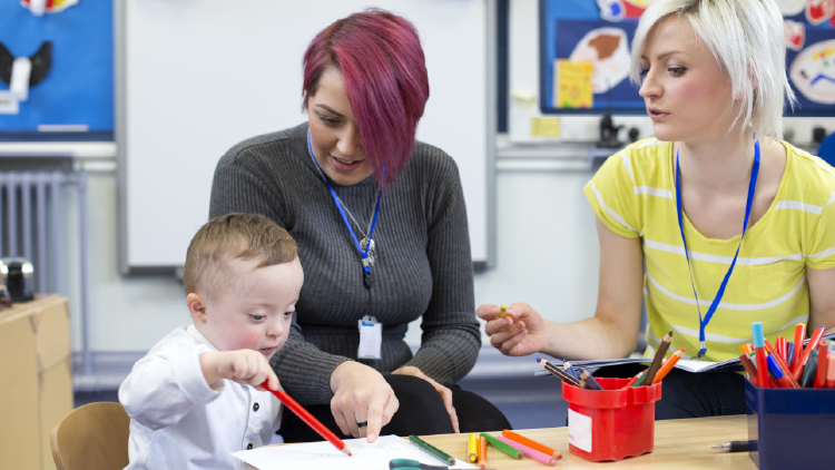 Two women sitting at a table with a special needs child who is drawing on a piece of paper while the woman closest to the child is pointing at the paper