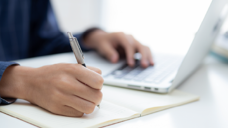 Hand writing on notepad with other hand in background typing on laptop
