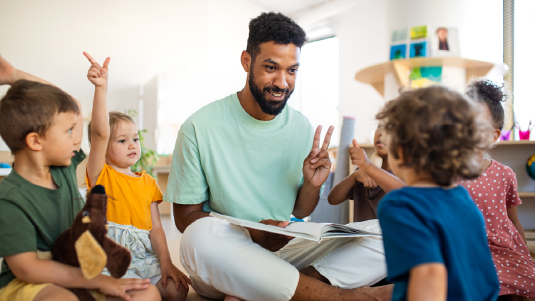 Man sitting crossed legged on the floor with a book holding up two fingers on his left hand with five children sitting on the floor surrounding him with their hands up