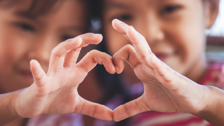 Close up of two young children's hands making a heart sign, their faces in the background