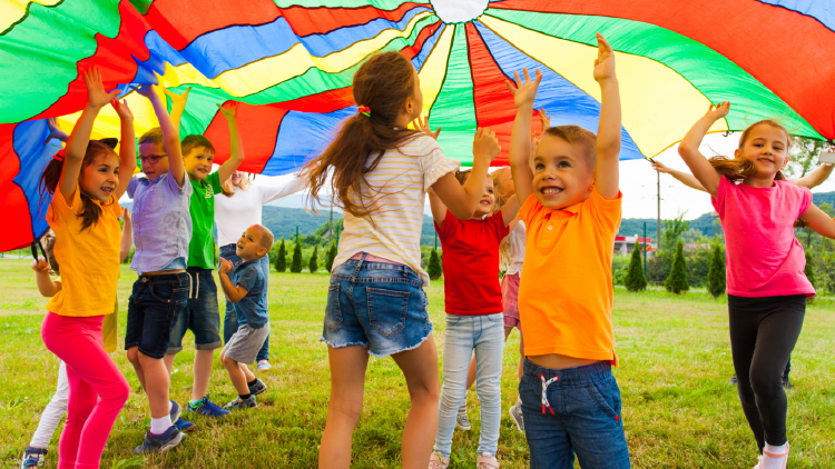 Several young people outside under a multicoloured parachute-like fabric that adults are floating above them