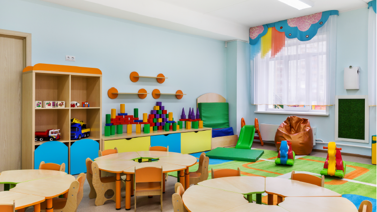 Wide view of childcare facility with round tables with chairs for young children, building blocks on counters and other toys in the room