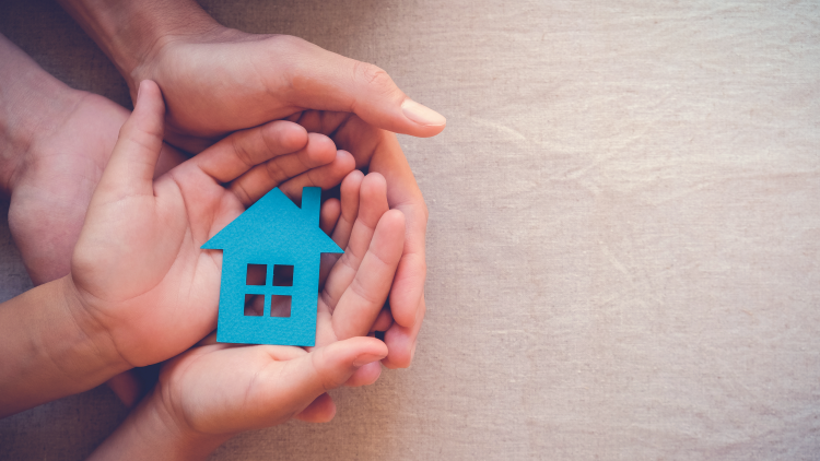 Close up of a young person holding a cut out of a blue house and an adult is then holding the young person's hands