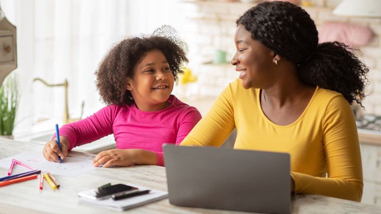 A woman and young child sit at a table. The woman sits with a laptop and the girl is holding a pencil, writing on paper. They're looking at each other and smiling.