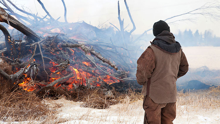 Person in a black touque, brown jacket and pants facing a pile of burning branches in a wintery field