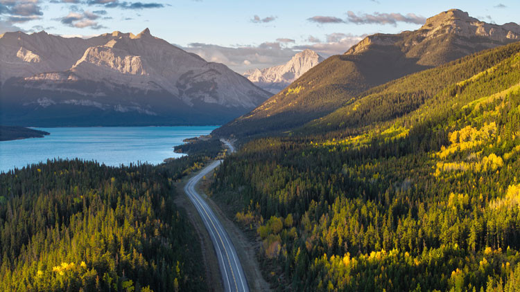 A winding road cuts throught the mountains with a lake on the left. The sun is shining through the clouds.
