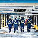 Four oil workers walk underneath a platform of pipes. They're wearing blue coveralls and white hard hats. There is snow on the ground.