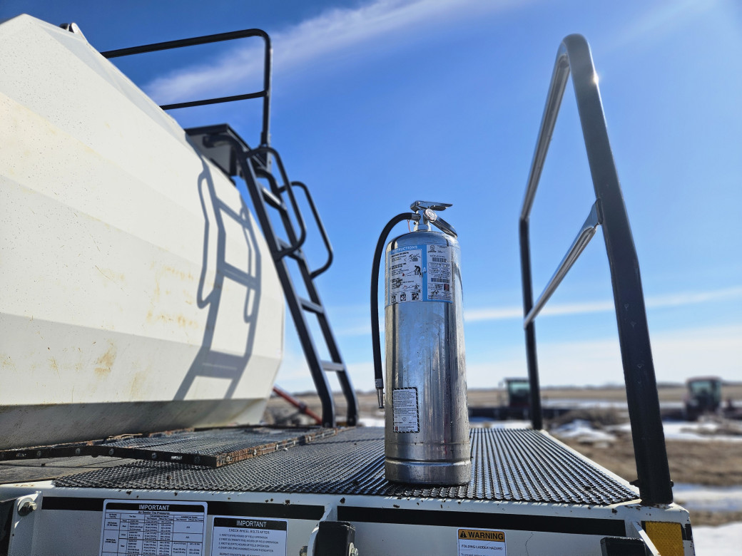 A tall, silver fire extinguisher sits on the platform of a large, white air seeder tank. In the background is a wide open field with other machinery. 