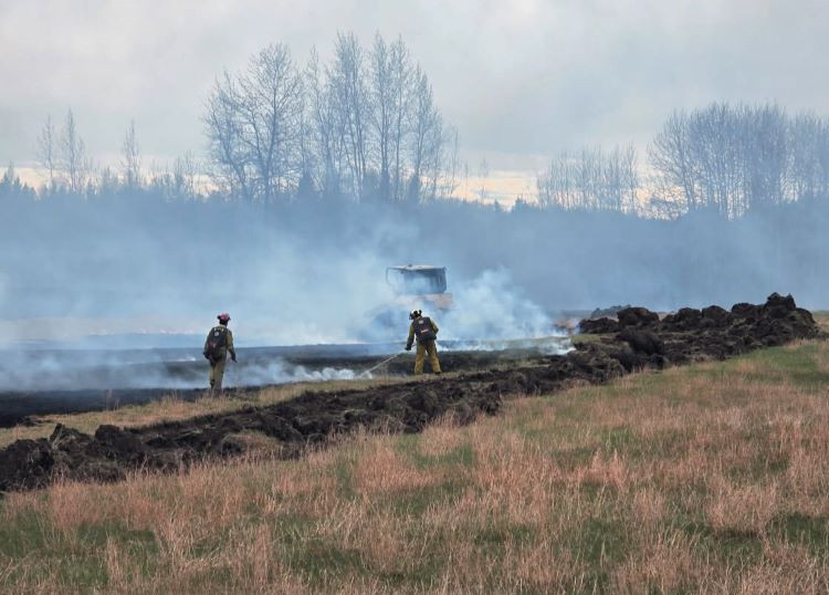 Firefighters in yellow shirts are containing a wildfire with wajax bags on their backs. The fire is burning in dead grass, creating a lot of smoke.