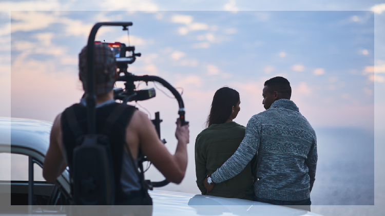 Cameraperson filming a couple standing beside a car at sunset, as the couple looks out over the water.”