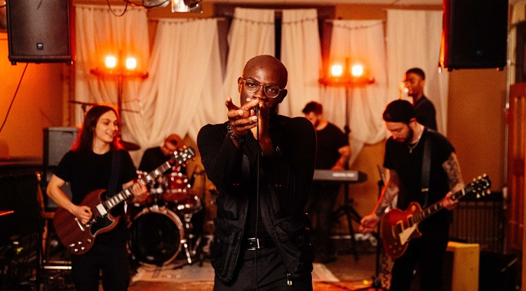 Performer sings into a microphone with his band behind him playing instruments.