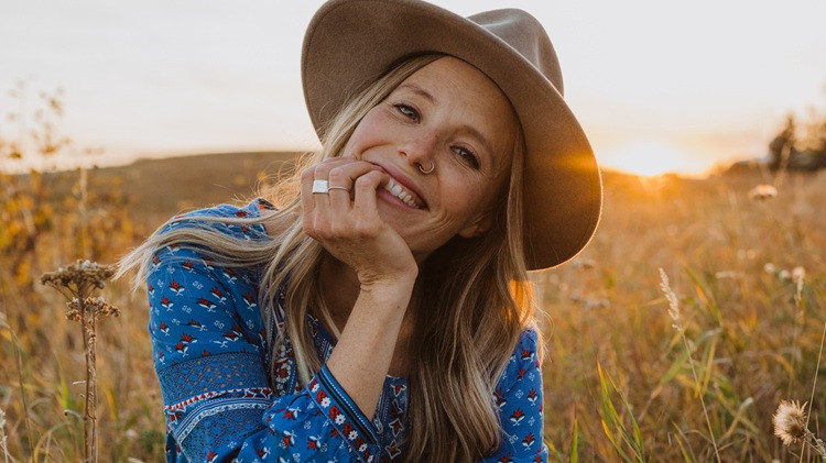 Person wearing a wide-brimmed hat and patterned blue shirt sitting in a grassy field at sunset, with the sun glowing in the background.