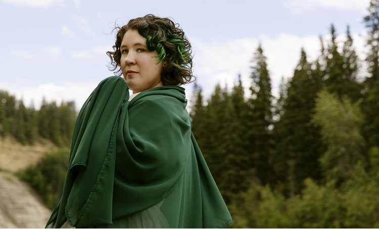 Performer is cloaked in a green shawl standing amongst evergreen trees.