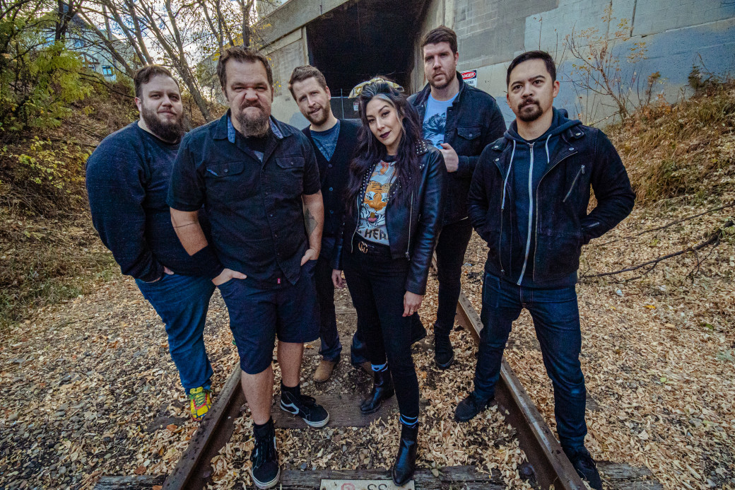Music band with 6 members stand together on train tracks near a bridge.