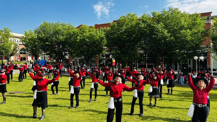 Dance group performs on an open green field. They have on red tops and hold their arms outstretched to the side.