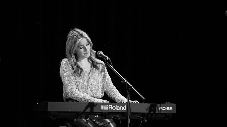 Black-and-white photo of a performer playing a keyboard and singing into a microphone on a dark stage.