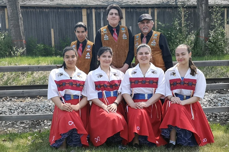 Dance group in traditional Metis clothing pose together in front of train tracks and a fence.